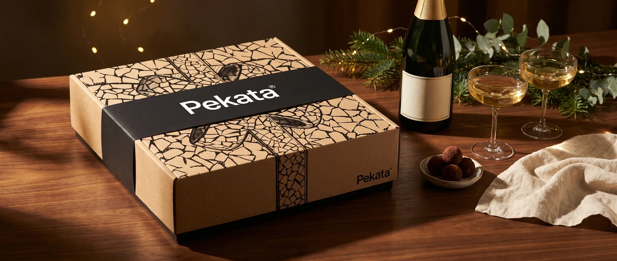 Closed Pekata Christmas box with a black band next to a bottle of cava and two glasses on a walnut table